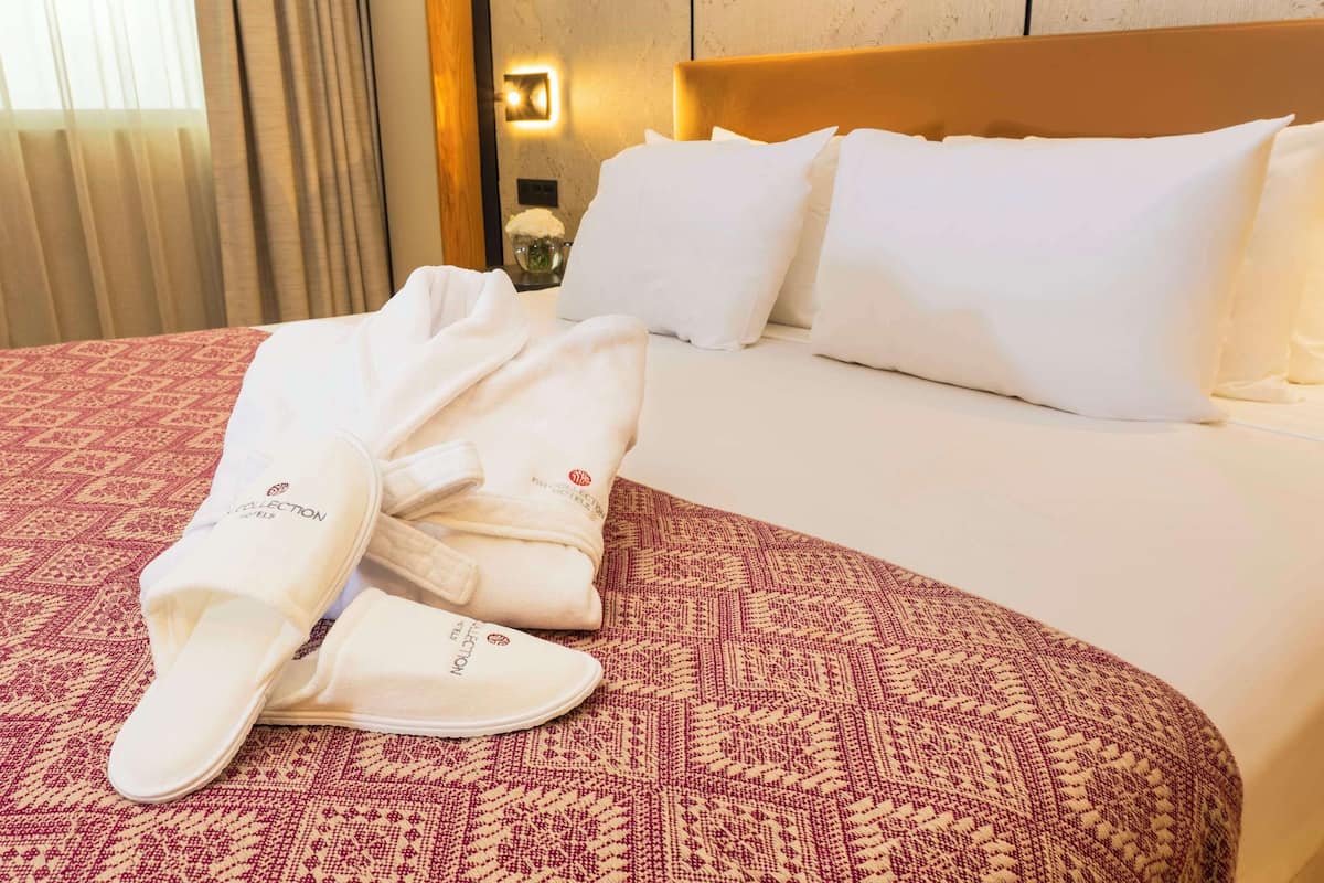 A hotel room with a bed, white pillows, a patterned red and beige bedspread, and a white robe with slippers on the bed.