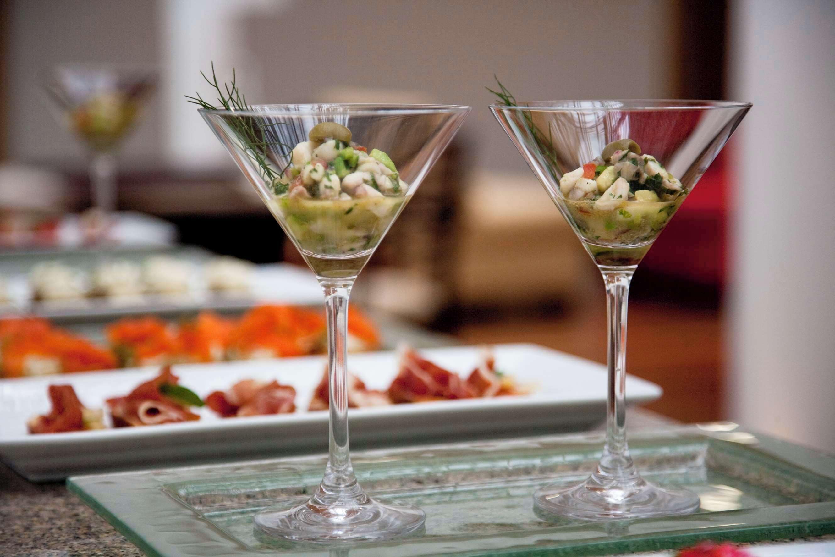 Deux verres à martini remplis d’une salade de fruits de mer, posés sur un plateau en verre, avec un arrière-plan flou composé d’assiettes remplies de nourriture.