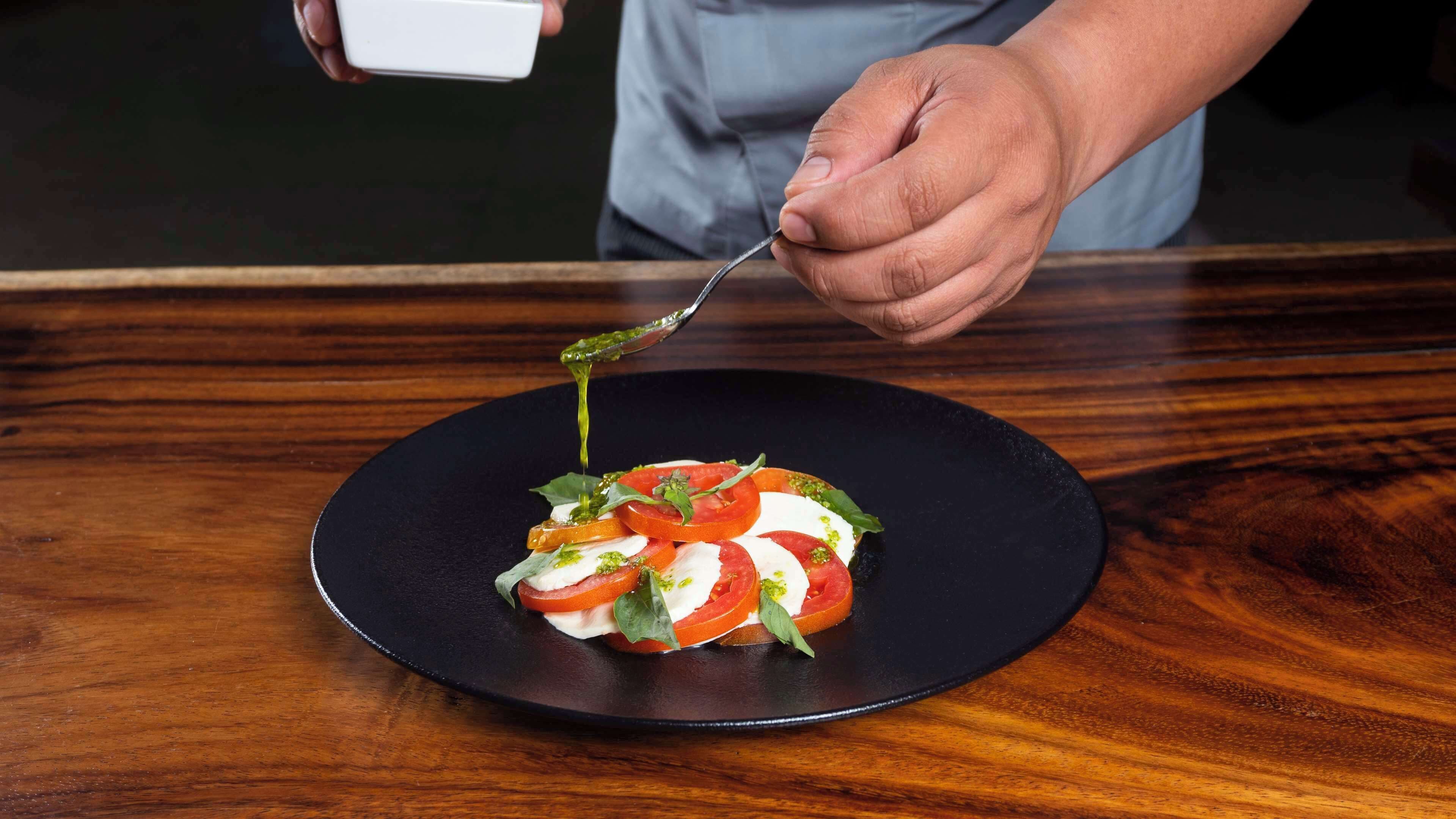 Un chef sirviendo salsa sobre un plato con tomate, mozzarella y albahaca sobre un plato negro.