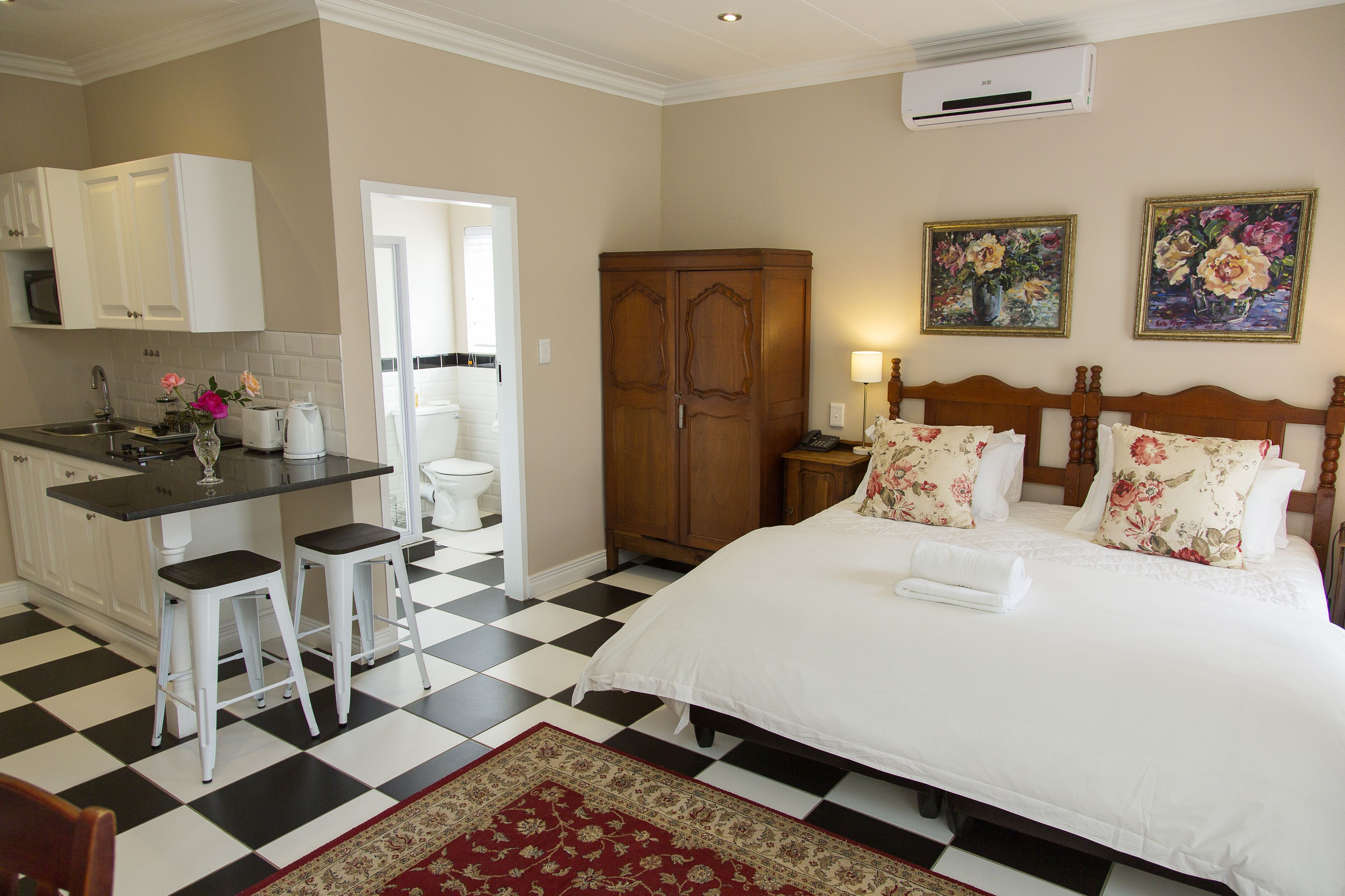 Una habitación de hotel con piso a cuadros, una cama con almohadas estampadas, un armario de madera, una cocina con gabinetes blancos y una alfombra roja.