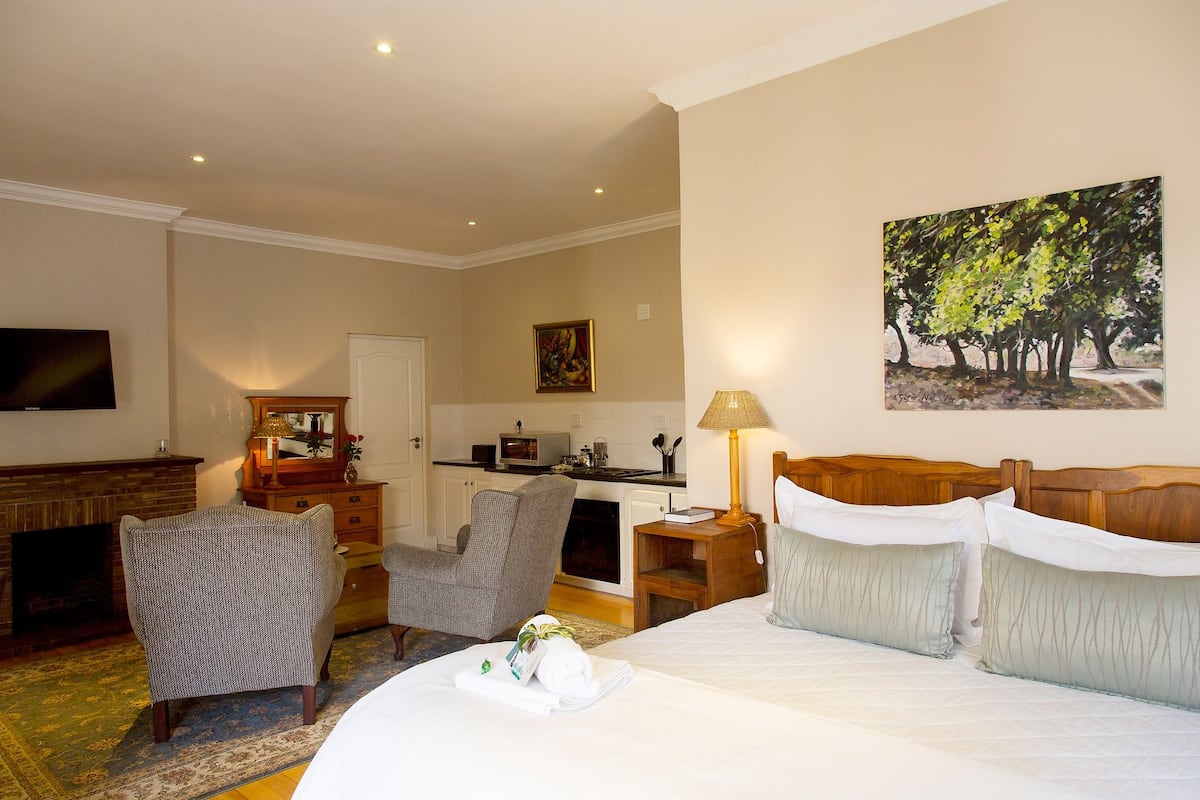A hotel room with a bed, a desk, a chair, a TV, a painting, and a kitchen area.