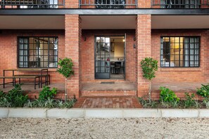 Un edificio de ladrillo de dos pisos con un balcón, un comedor con mesa y sillas, y un pequeño jardín con plantas.