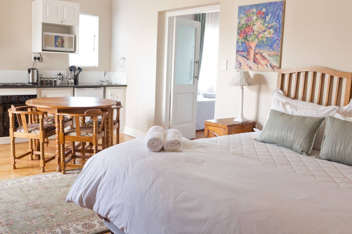 A hotel room with a bed, a wooden dining table, chairs, a kitchen area, and a painting on the wall.