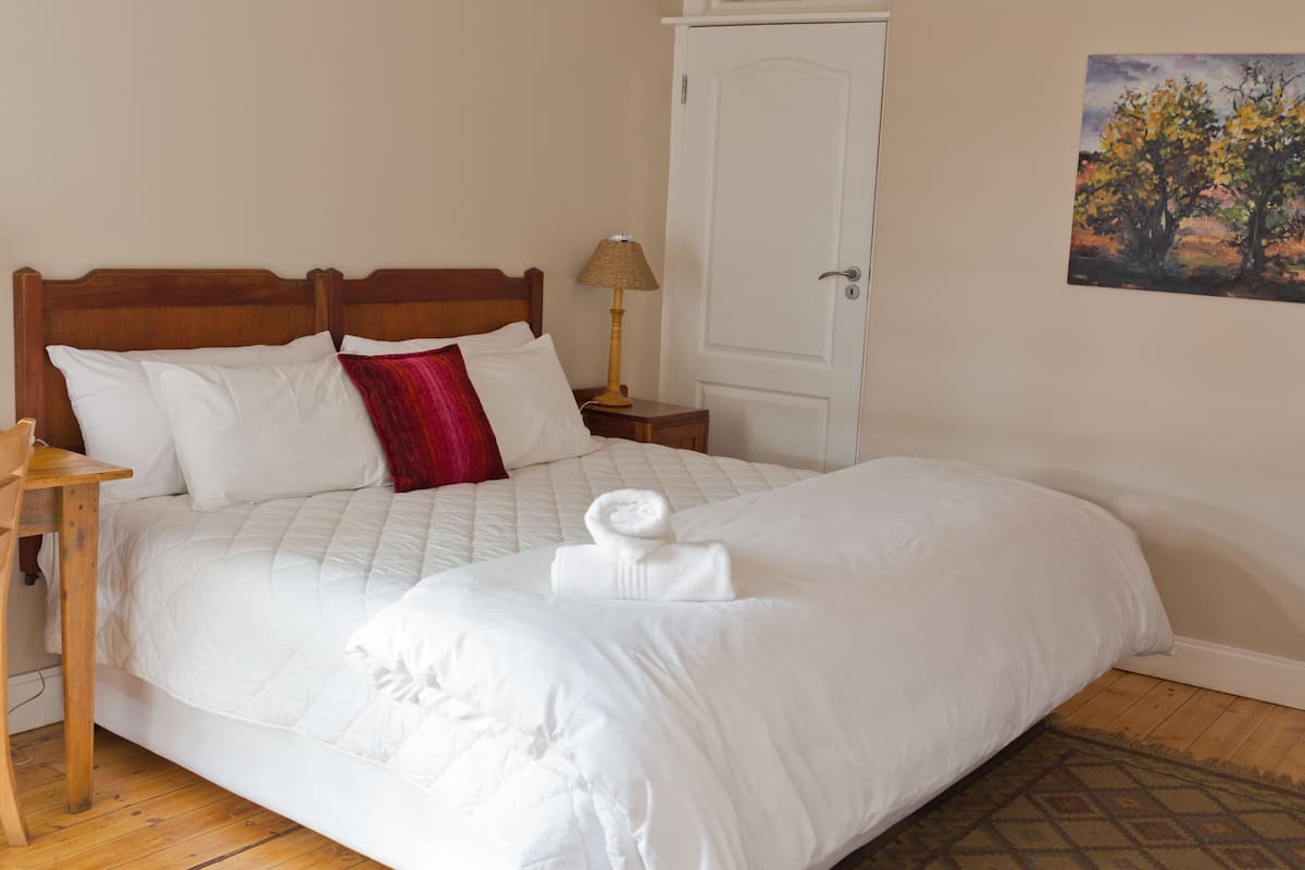 A neatly made bed with white linens and a red throw pillow, a wooden nightstand with a lamp, and a door with a painting of a tree on the wall.
