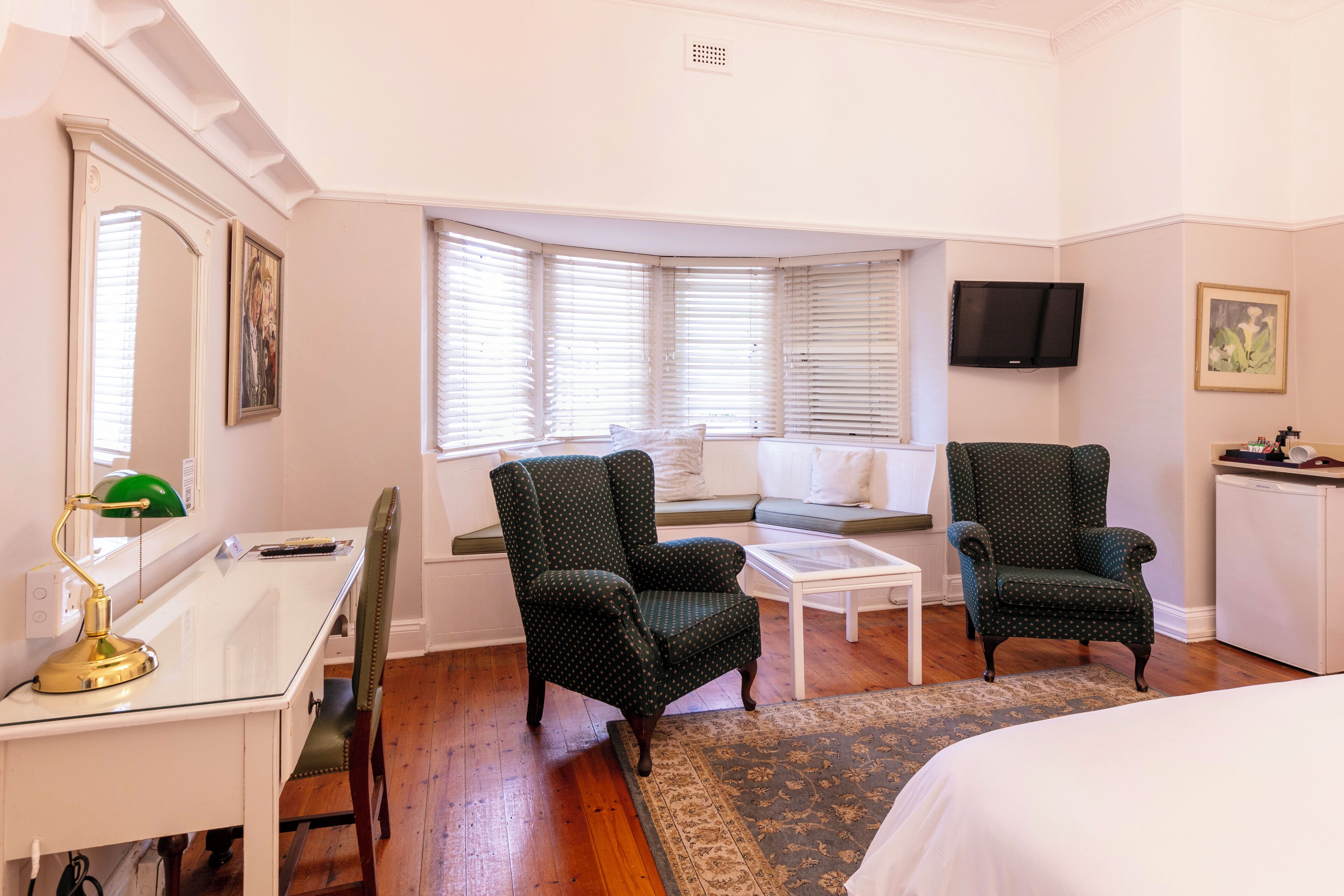 A hotel room with a desk, two armchairs, a bed, a window seat, and a television.