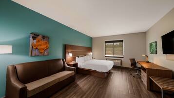 A hotel room with a bed, a wooden desk, a brown leather sofa, a nightstand, and a window with a view of trees.