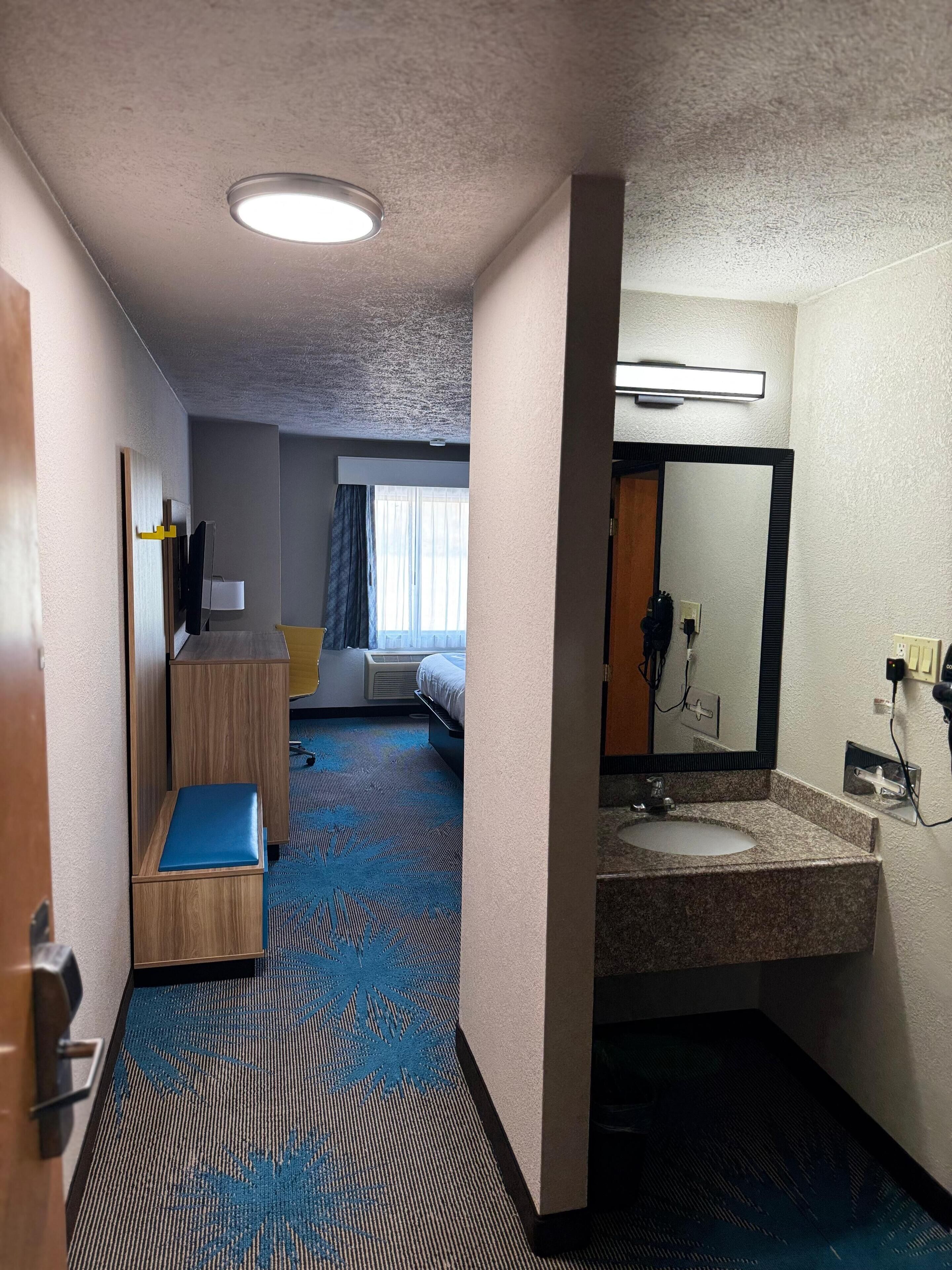Habitación de hotel con cama, escritorio, silla, baño con lavabo y espejo, y piso alfombrado.