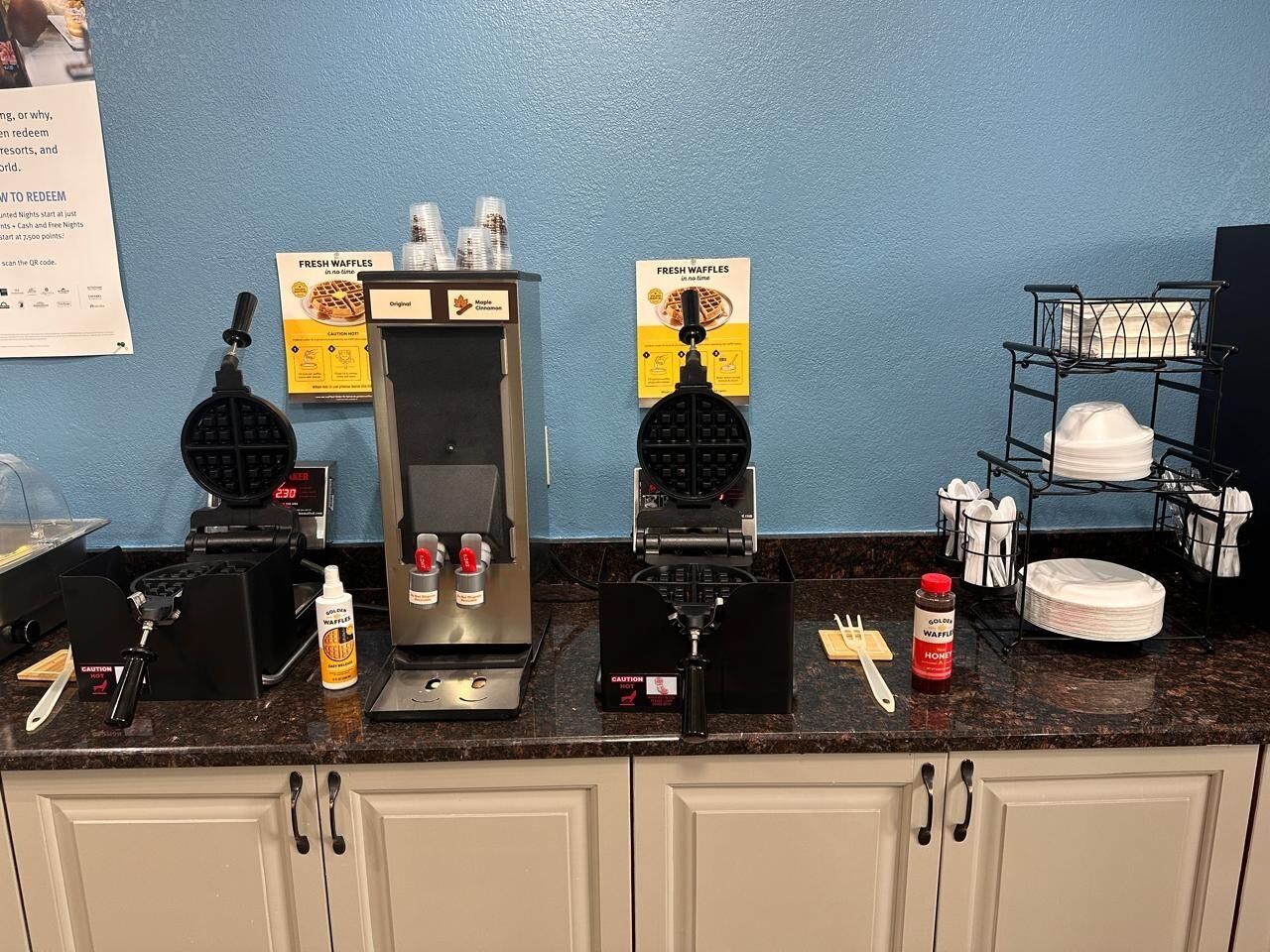 A self-service breakfast station with waffle makers, condiments, and a coffee machine.