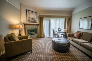 A hotel room with a patterned carpet, a sofa, a chair, a dining table with chairs, a fireplace, and a large window with curtains.