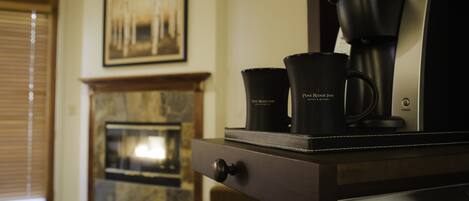 Una cafetera con dos tazas negras sobre una mesa de madera, una chimenea y un cuadro en la pared.
