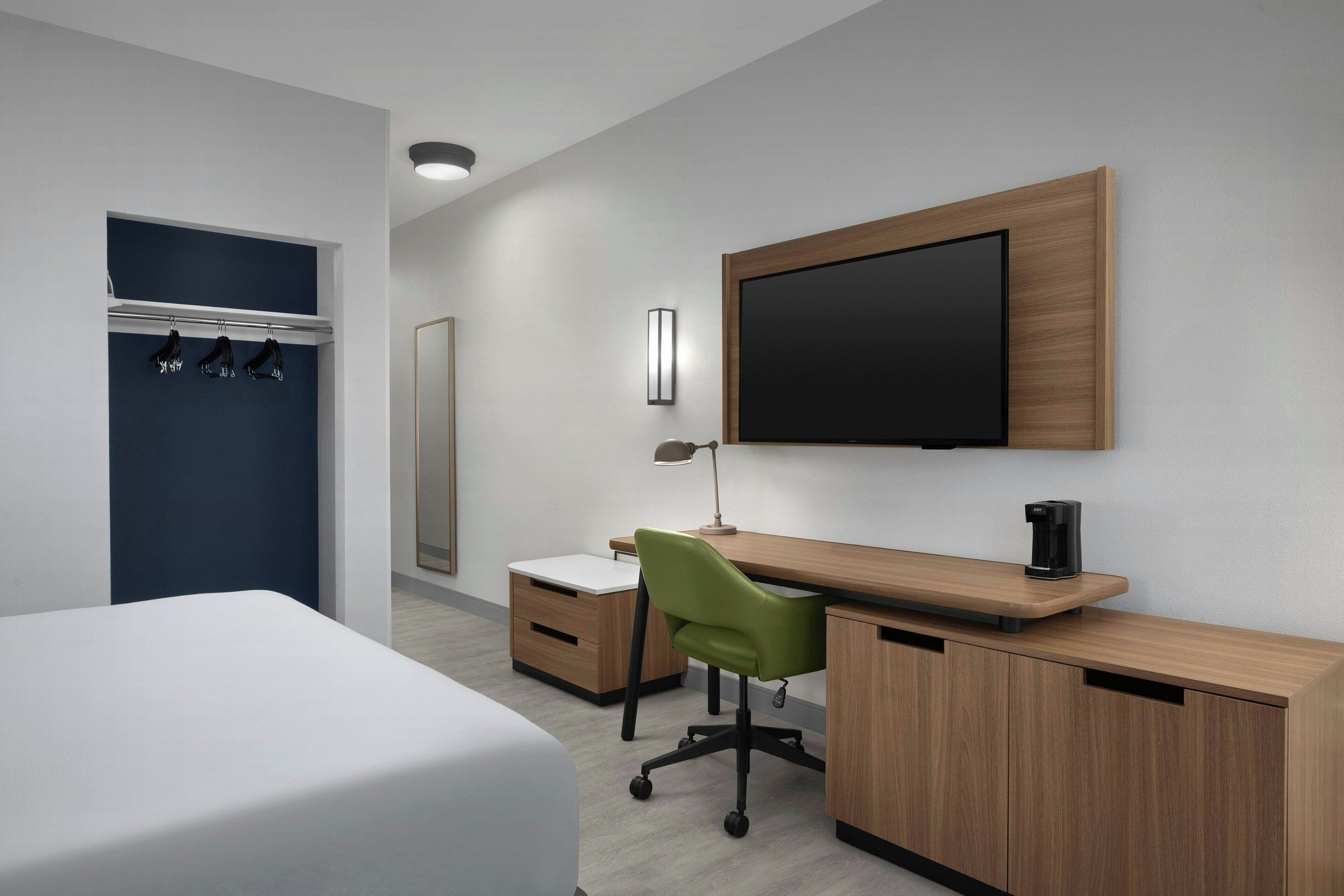 A modern hotel room with a bed, a desk with a chair, a TV mounted on the wall, and a coat rack.