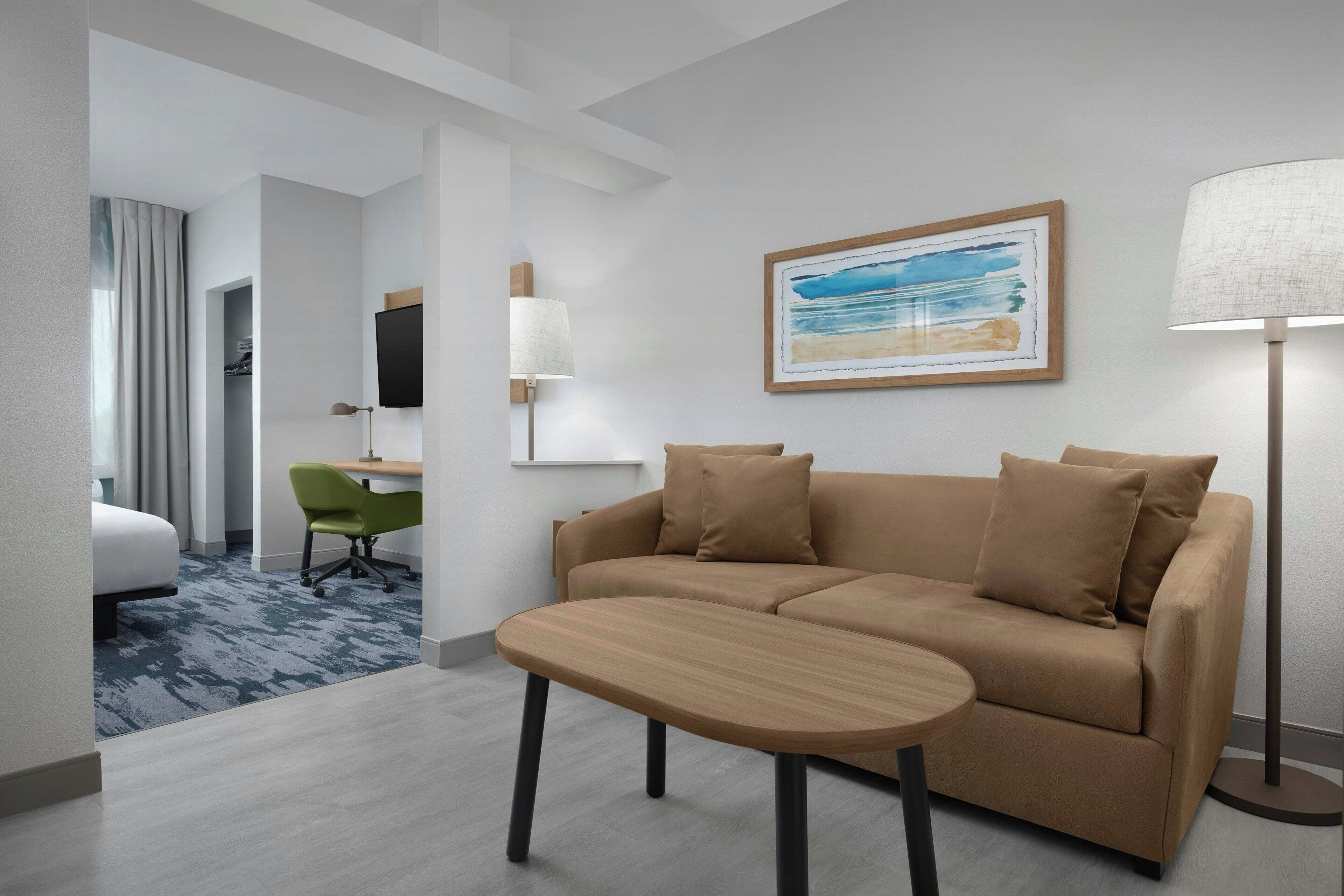 Une chambre d’hôtel moderne avec un canapé beige, une petite table ronde, un fauteuil de bureau vert, un téléviseur fixé au mur et une lampe sur pied.