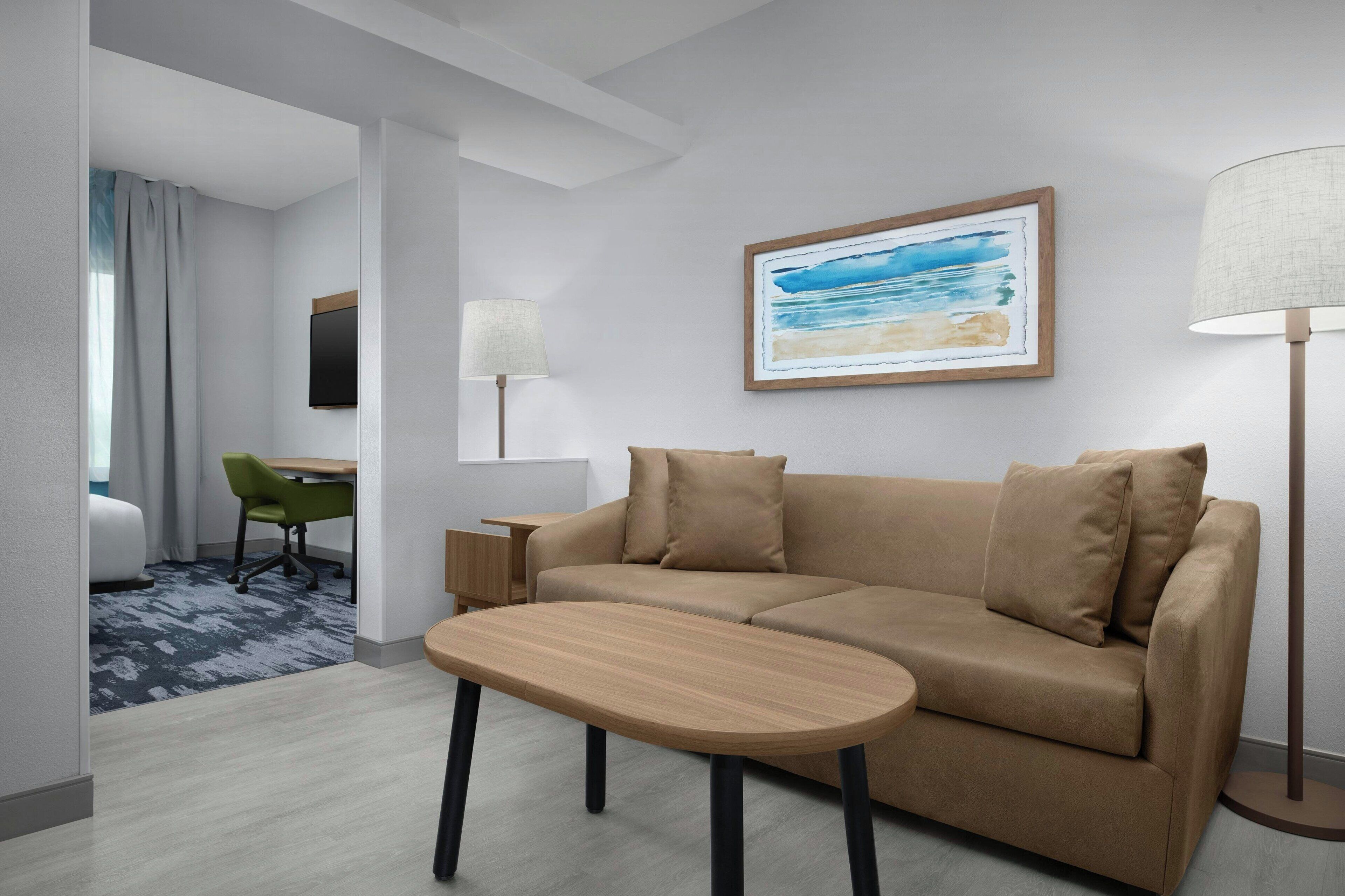 Un moderno cuarto de hotel con un sofá color beige, una pequeña mesa de madera, una silla verde, un escritorio, una lámpara y un cuadro enmarcado con una escena de playa.