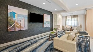 Un moderno vestíbulo de hotel con una alfombra grande de diseño abstracto, un sofá color beige, un televisor de pantalla plana en la pared y una zona de comedor con mesa y sillas.