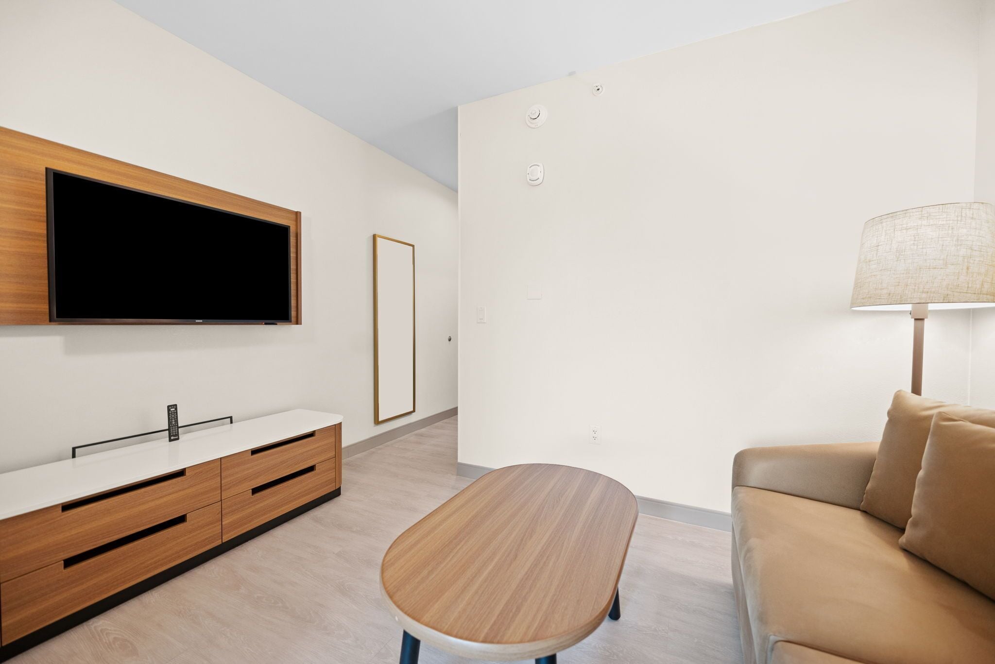 A modern living room with a flat-screen TV mounted on the wall, a wooden coffee table, a beige sofa, and a side table with a lamp.
