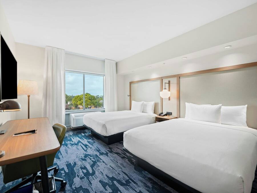 Une chambre d’hôtel avec deux lits, un bureau, une chaise, une télévision et une grande fenêtre donnant sur des arbres.