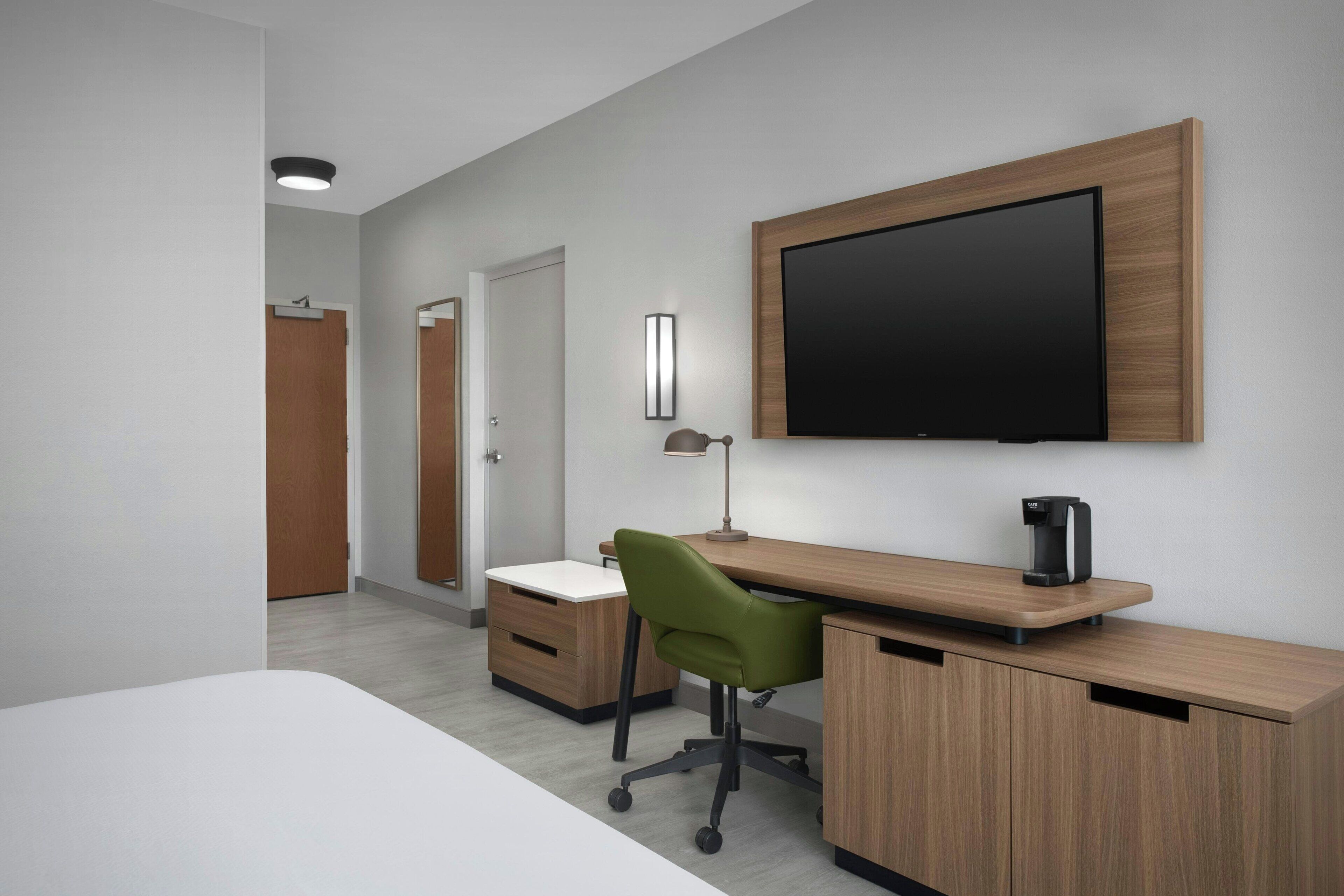 Quarto de hotel moderno com escrivaninha de madeira, cadeira de escritório verde e televisão de tela plana fixada na parede.