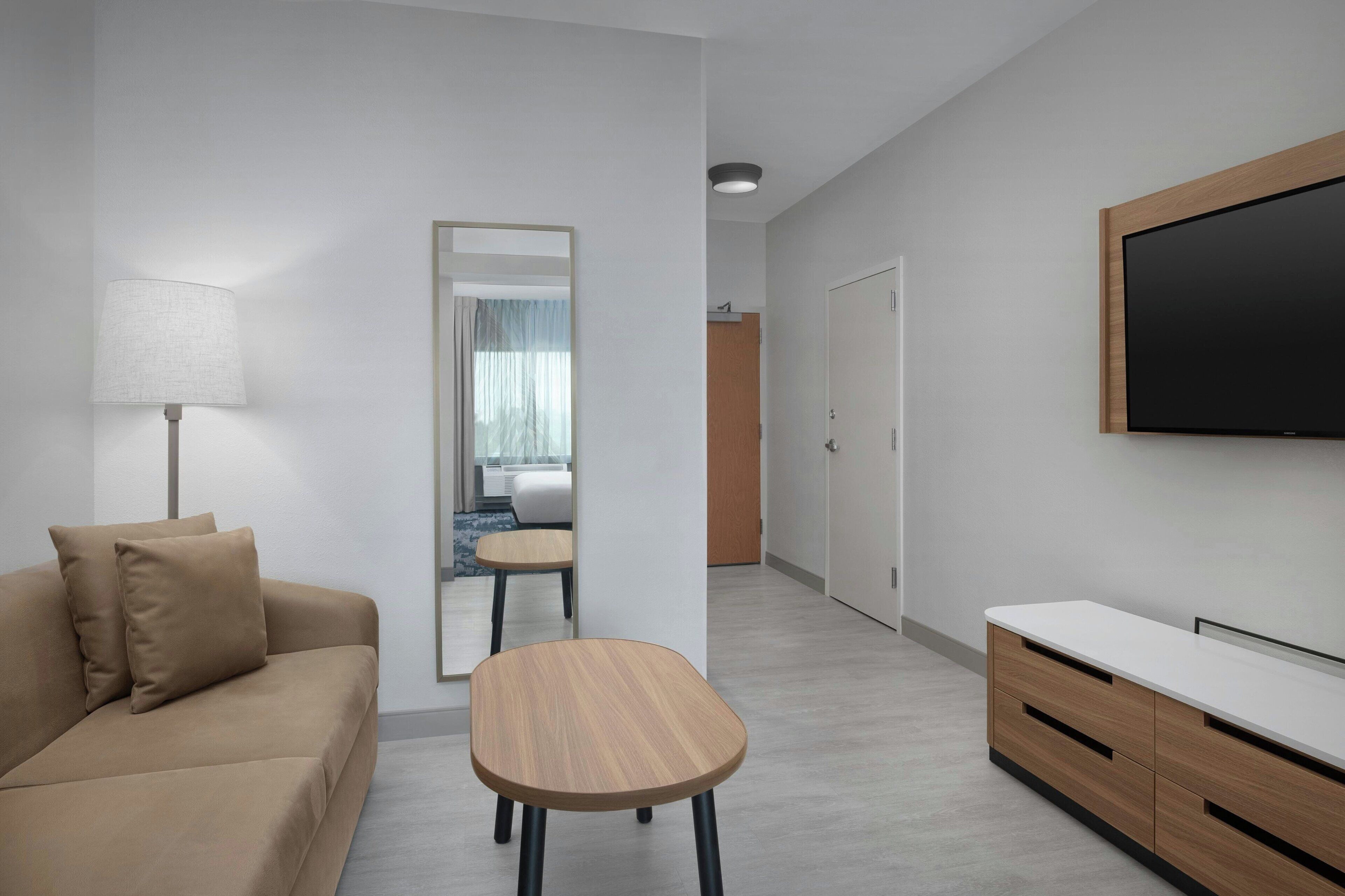 A modern hotel room with a sofa, a small wooden table, a TV mounted on the wall, and a mirror.