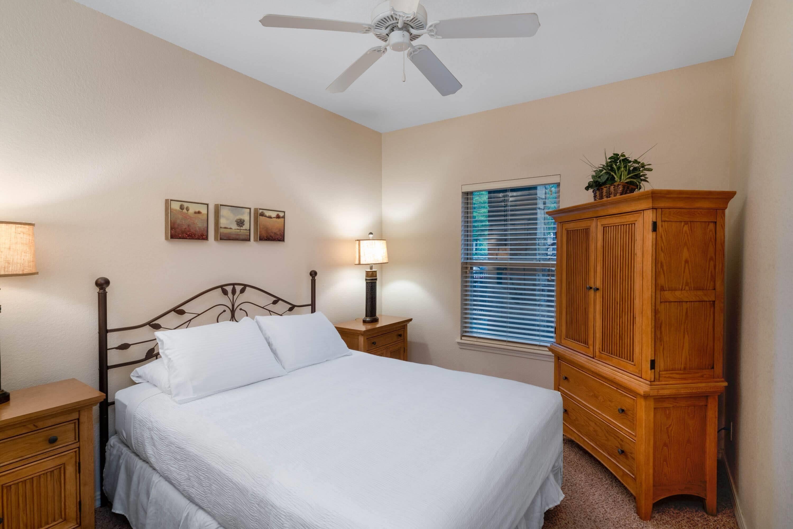 Une chambre à coucher comprenant un lit, une table de chevet en bois, une petite table de chevet avec une lampe, un ventilateur de plafond, une fenêtre avec des stores et une armoire en bois.