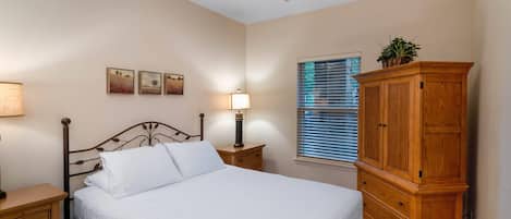 Une chambre à coucher comprenant un lit, une table de chevet en bois, une petite table de chevet avec une lampe, un ventilateur de plafond, une fenêtre avec des stores et une armoire en bois.