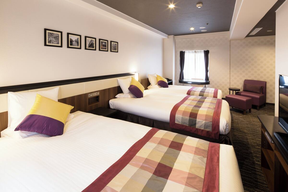 Habitación de hotel con tres camas, televisión, una mesita y un ventanal con cortinas.