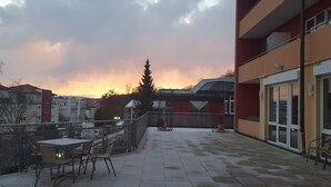 Una terraza en la azotea con vistas a la puesta de sol, mobiliario exterior y un edificio de fachada roja.