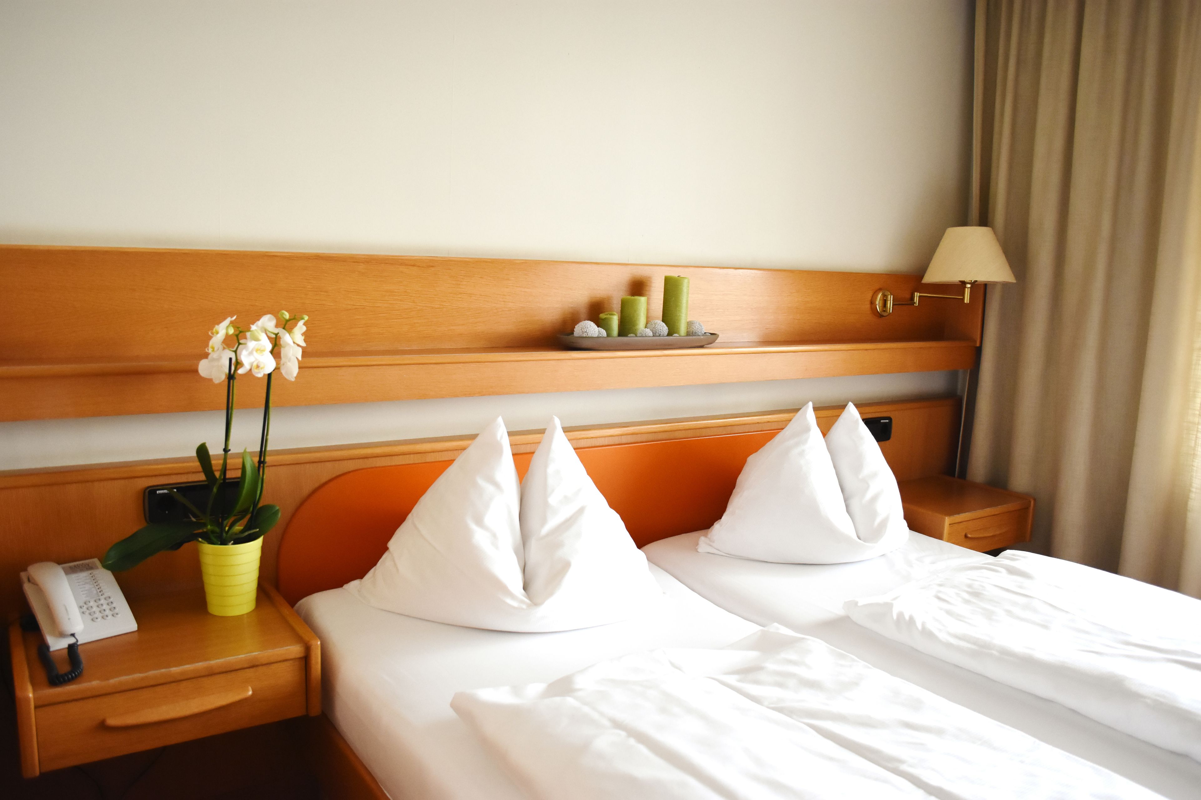 A hotel room with a wooden headboard, a bedside table with a lamp, a phone, a plant, and a tray with candles.