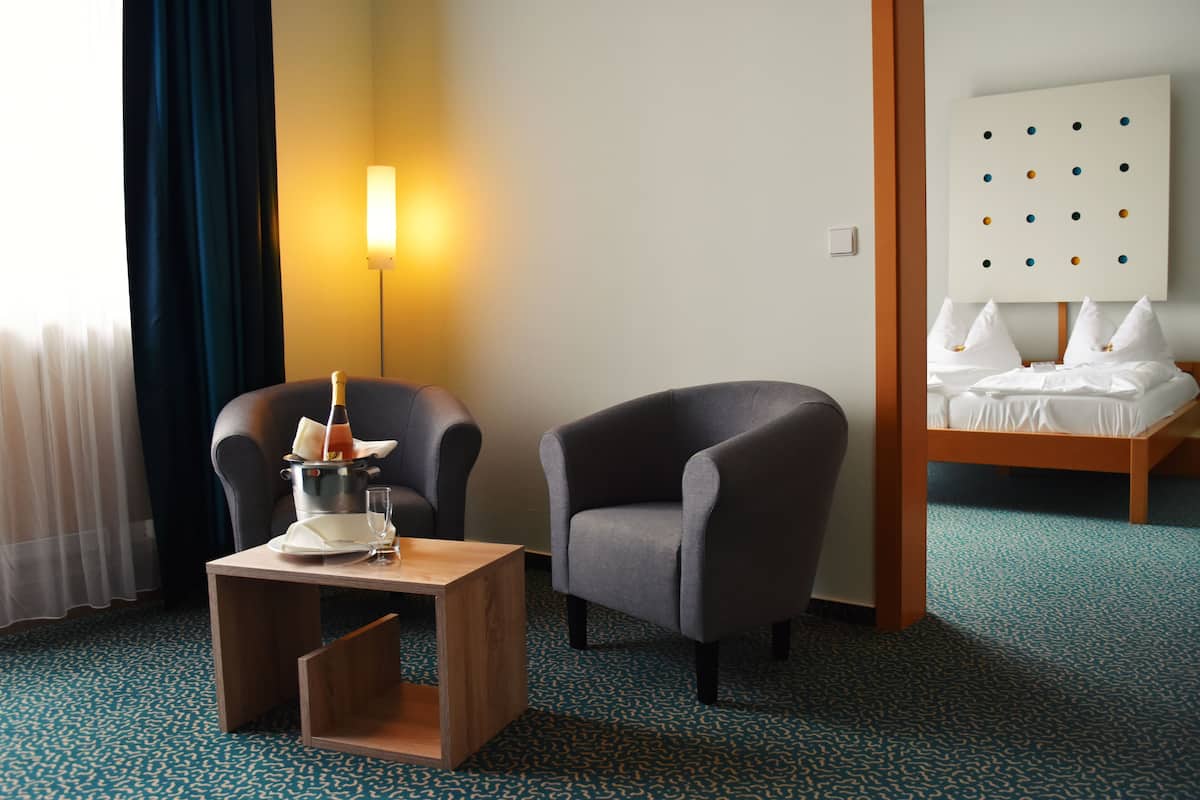 A hotel room with a bed, two armchairs, a small wooden table with a bottle and glasses, a lamp, and a wall with a dot pattern.