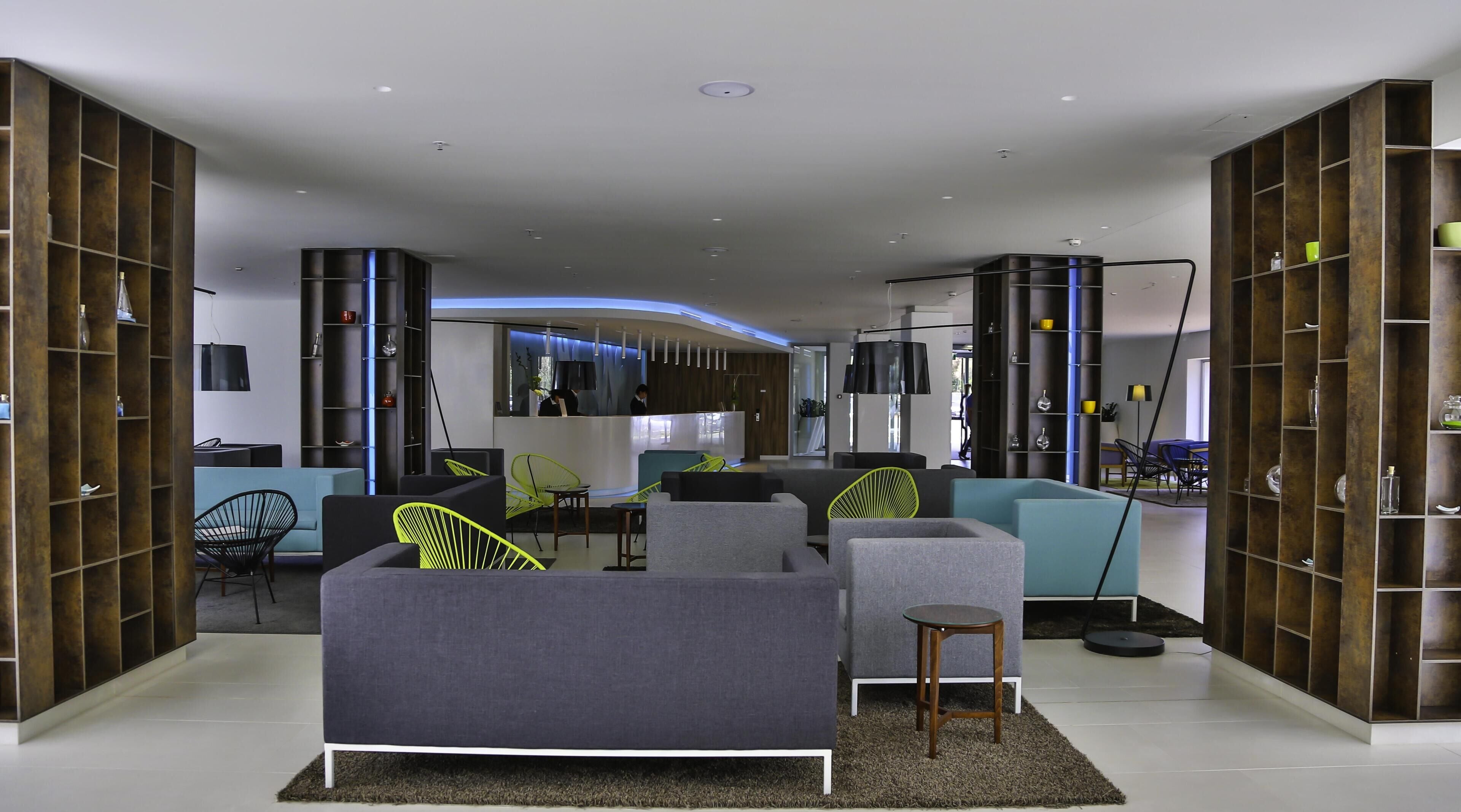 A modern living room with a grey sofa, a small wooden table, and a seating area with chairs. There are bookshelves on either side of the room.