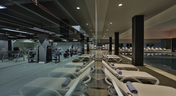 Un gimnasio moderno con equipo de ejercicio, un pasillo central y sillones de descanso con toallas.