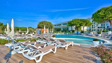 A poolside area with sun loungers and umbrellas, surrounded by greenery and a modern building.
