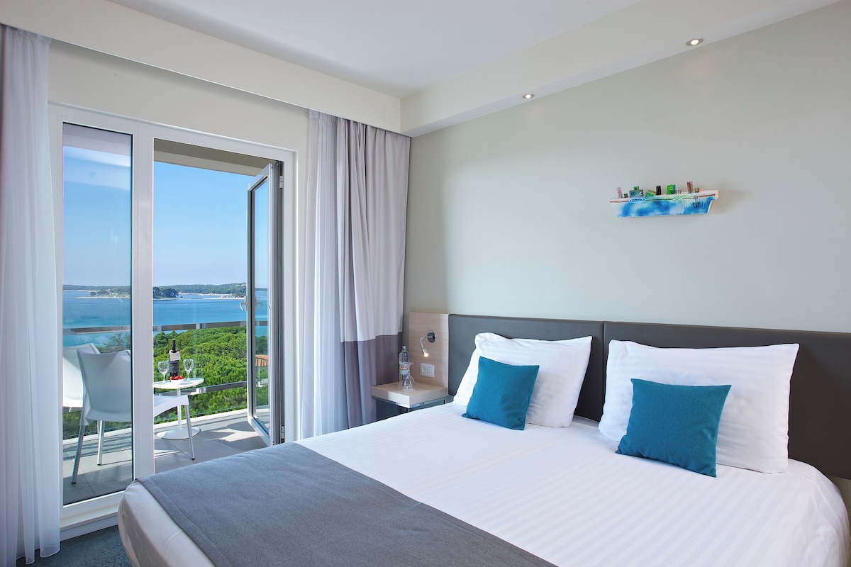 Habitación de hotel con una cama grande, un balcón con vistas al mar, una mesita con una botella y un vaso, y una mesita de noche con una lámpara.