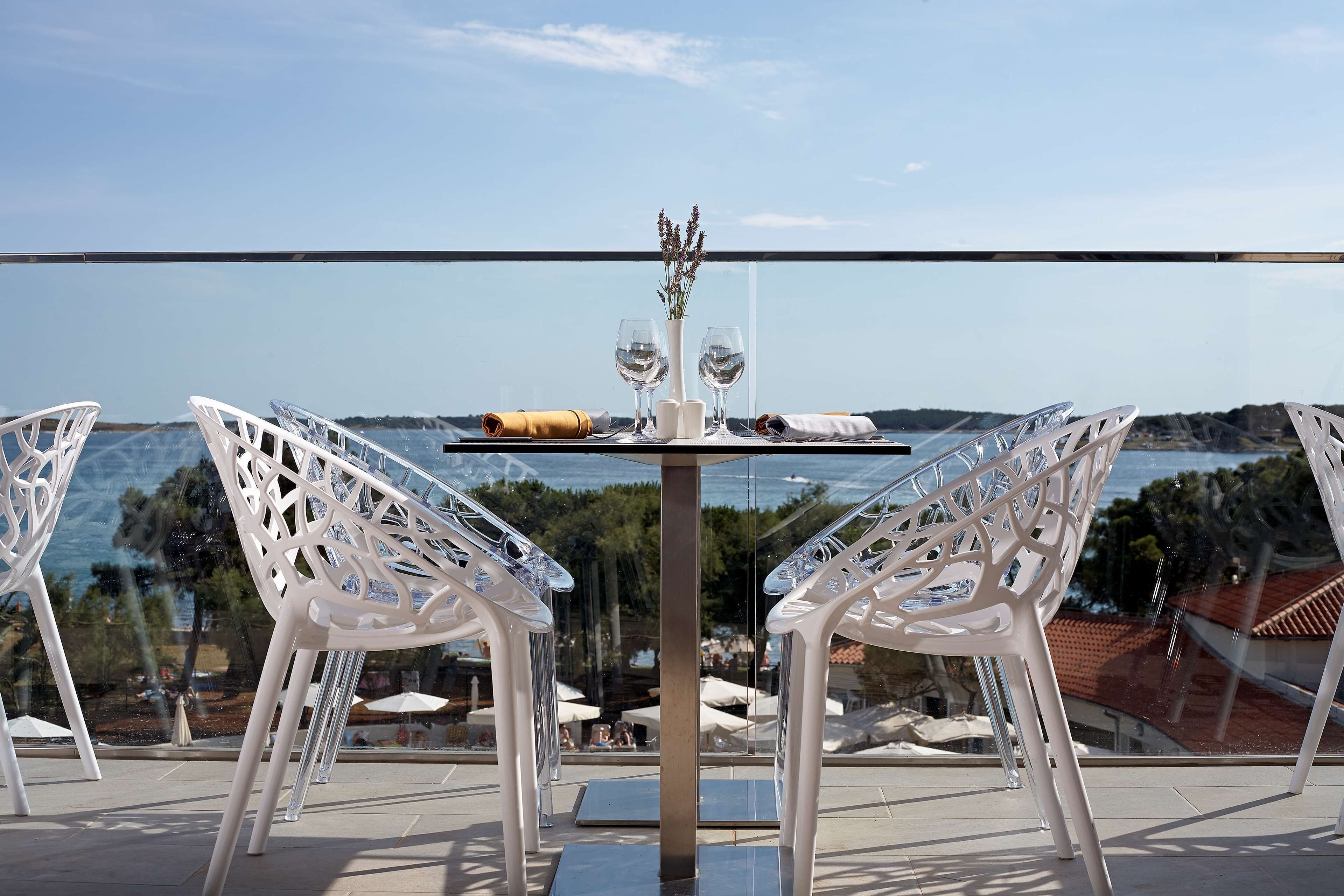 A table with two chairs, a vase with flowers, and two wine glasses set on a balcony overlooking a body of water and a distant landscape.