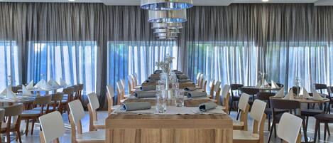 A large dining area with multiple tables and chairs, a central island with a vase of flowers, and large windows with curtains.