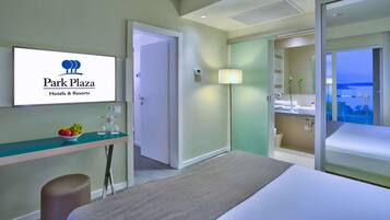 A hotel room with a bed, a desk with a bowl of fruit and glasses, a bathroom with a sink and mirror, and a view of the outdoors.
