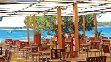 Café al aire libre con mesas y sillas de madera, con vistas a una playa y al mar.