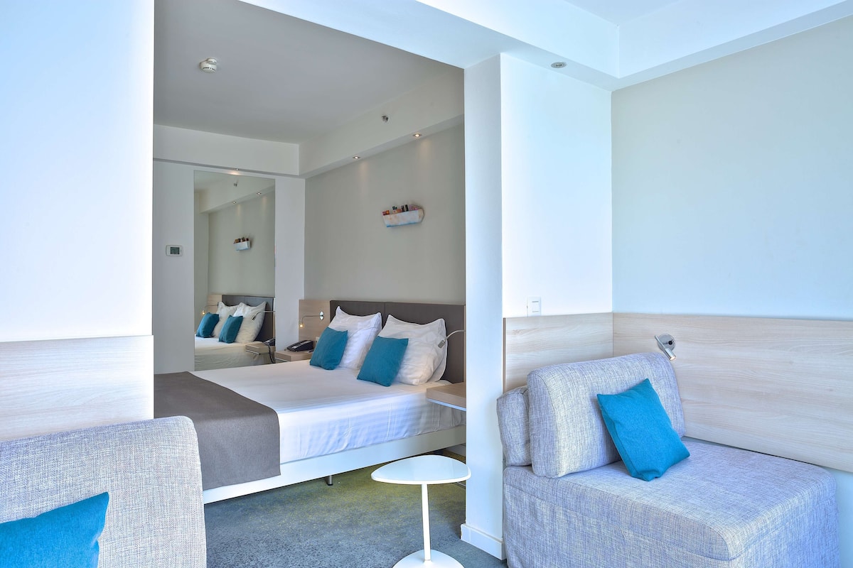 A modern hotel room with a large bed, a sofa, a small table, and a mirror reflecting the room.