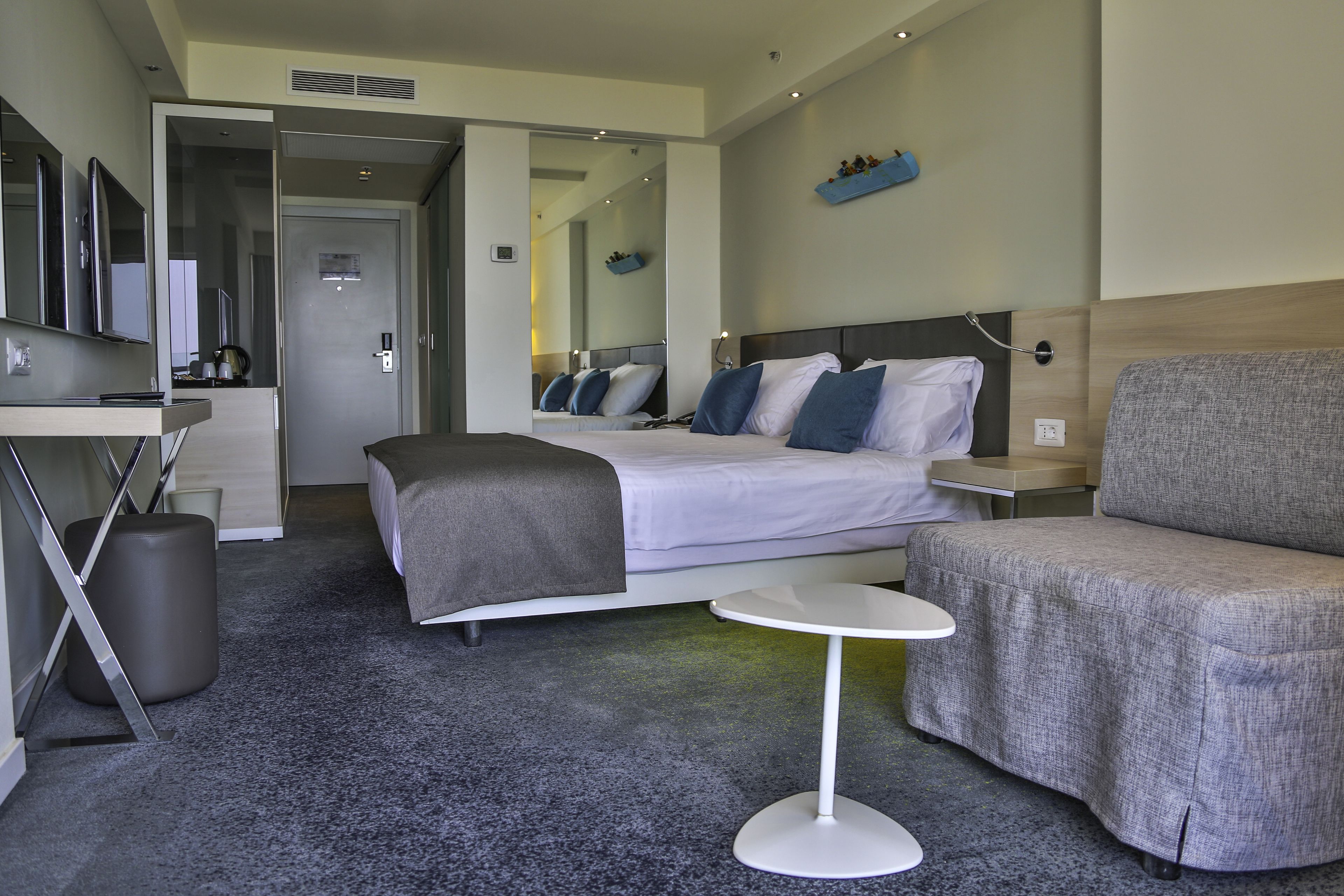 A modern hotel room with a bed, a grey sofa, a small white table, and a TV on a stand.