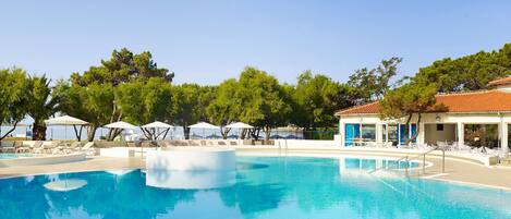 A clear blue swimming pool with lounge chairs and umbrellas, surrounded by trees and a building with a tiled roof.