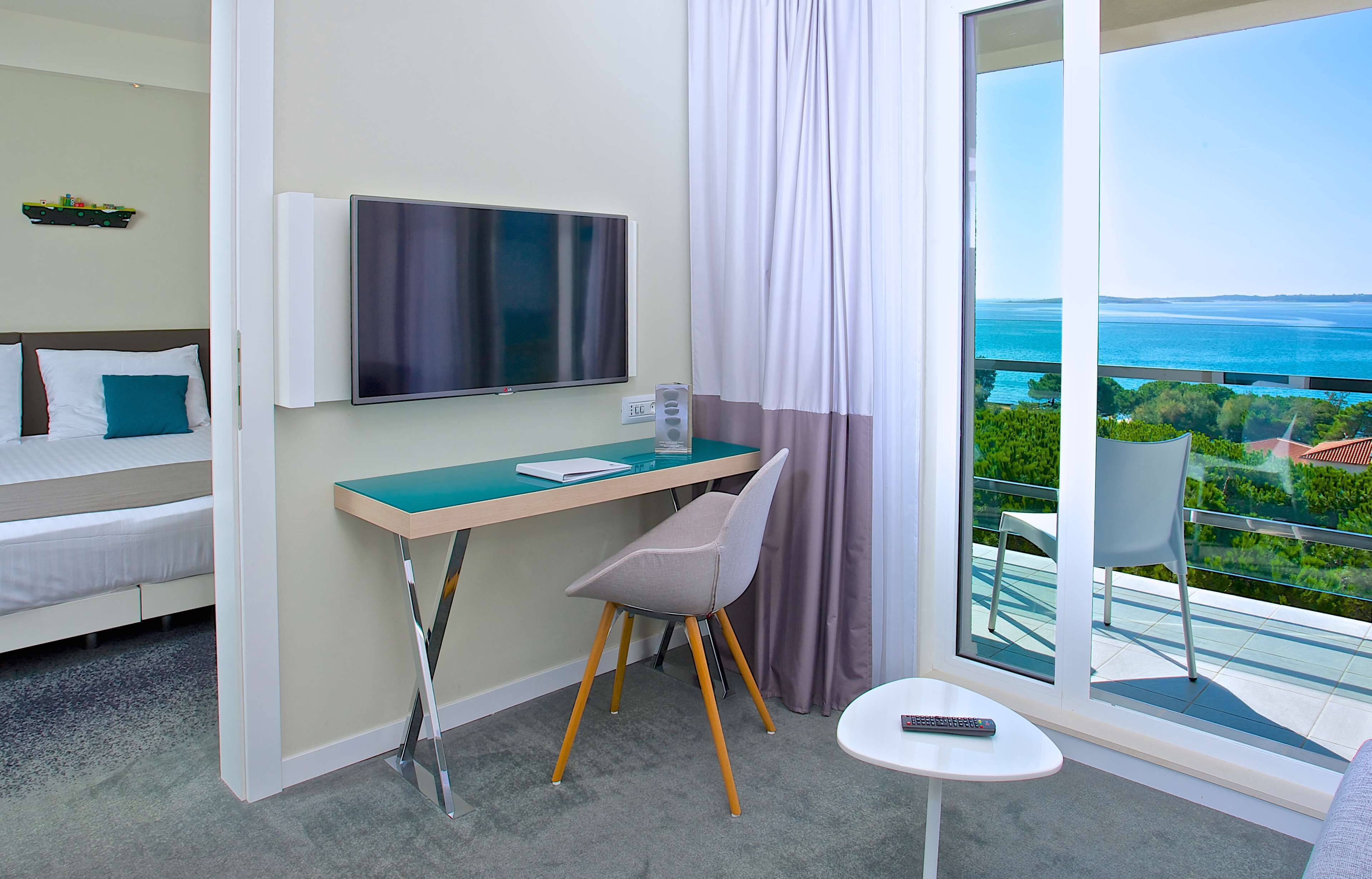A hotel room with a bed, a desk with a chair, a TV, a small table, and a balcony with a view of the sea.