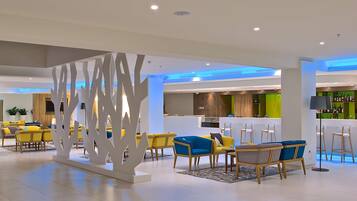 Eine moderne Hotellobby mit gelb-blauen Sitzgelegenheiten, einem Barbereich und einer Küche im Hintergrund.