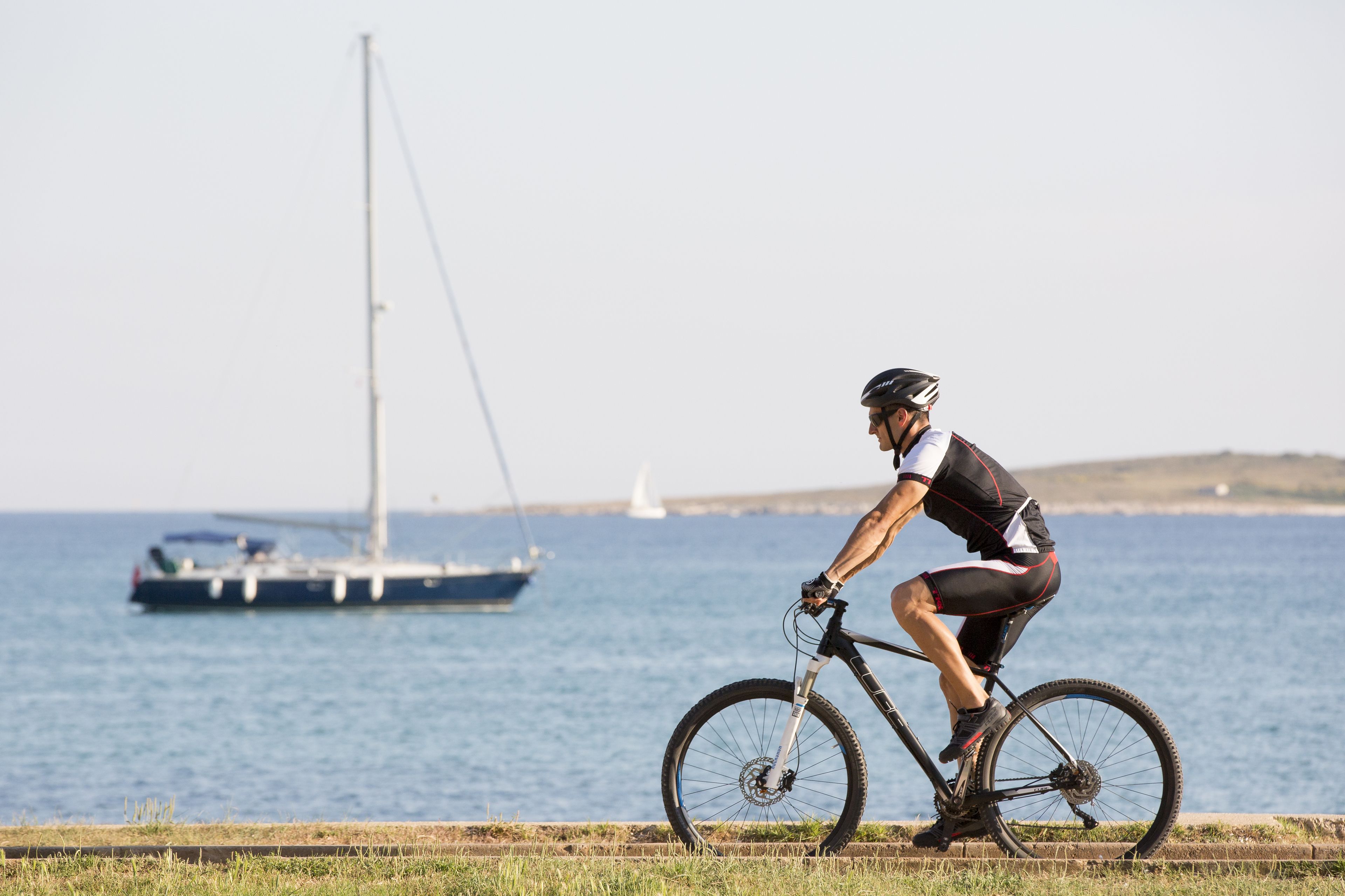 A cyclist riding a bike by the sea with a sailboat in the background.