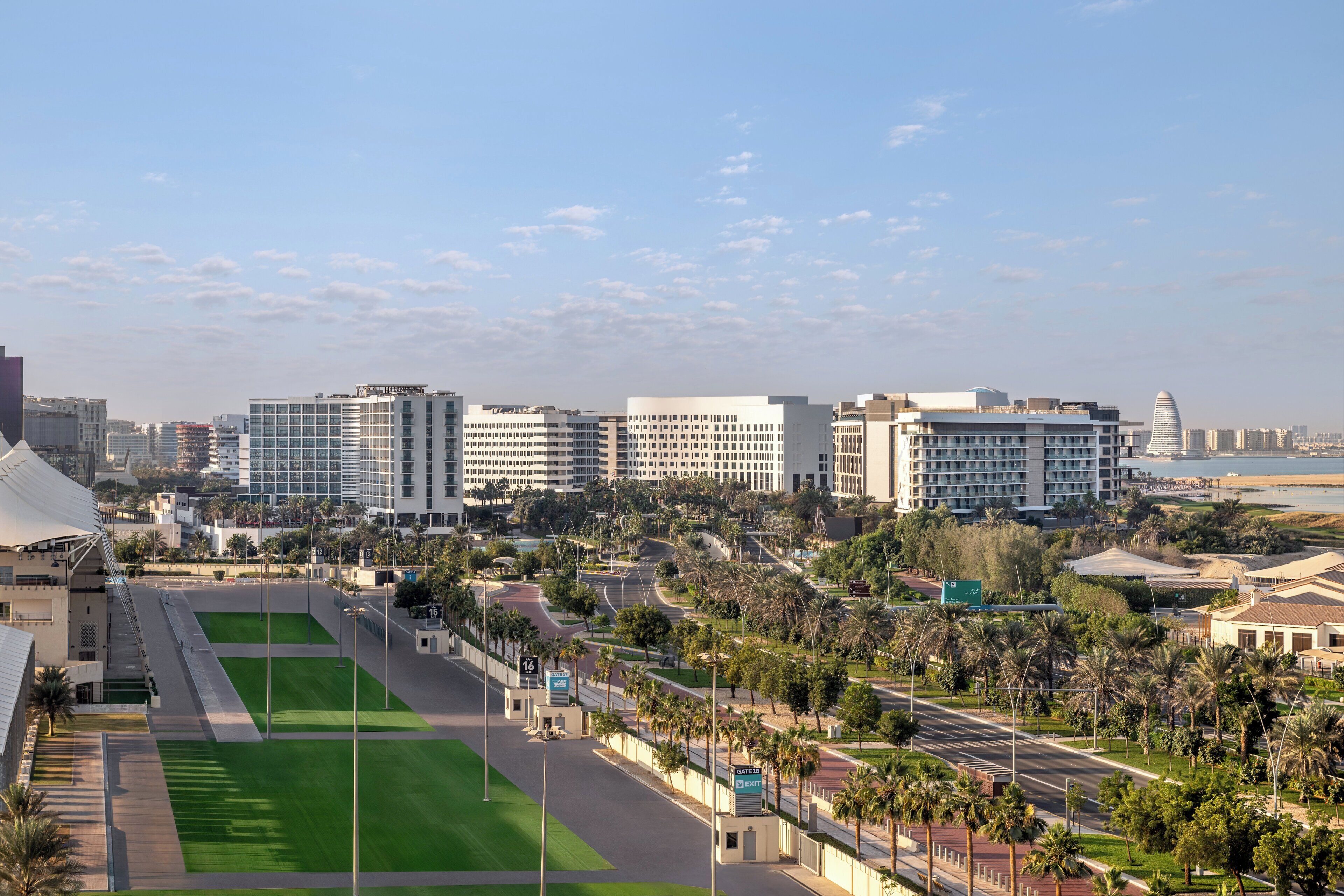 A cityscape with modern buildings, a green sports field, and a coastal view.