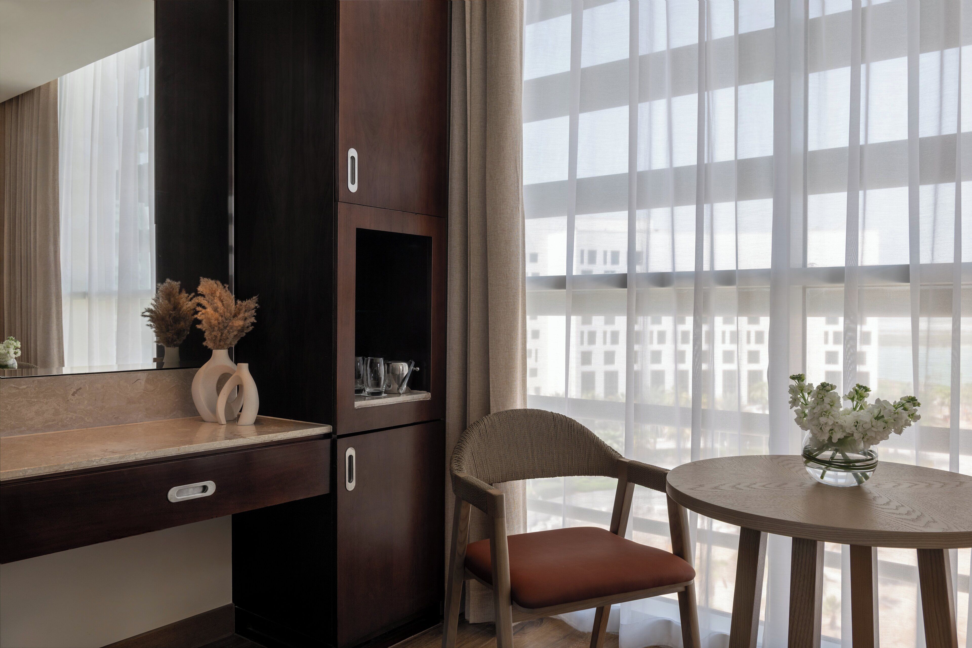 A modern hotel room with a large window, a wooden chair, a small round table with a vase of flowers, and a minibar.