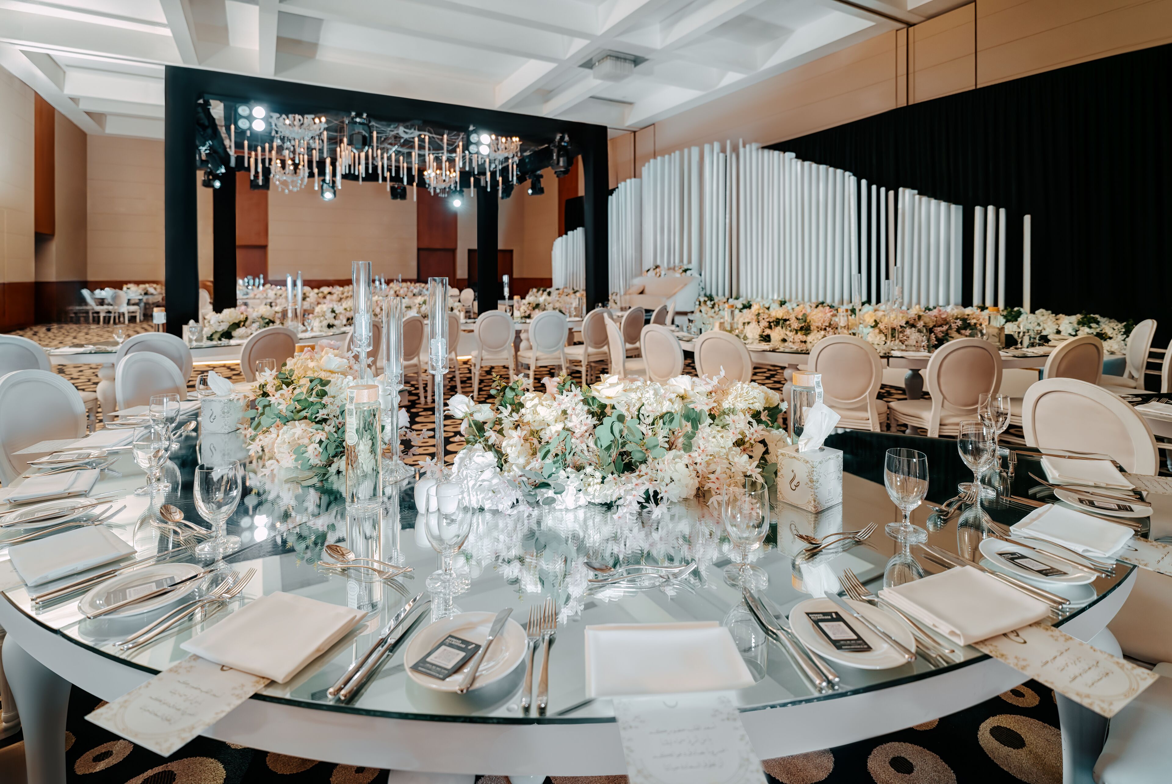 A banquet hall with round tables set for a formal event, featuring glass tops, white tablecloths, and floral centerpieces.