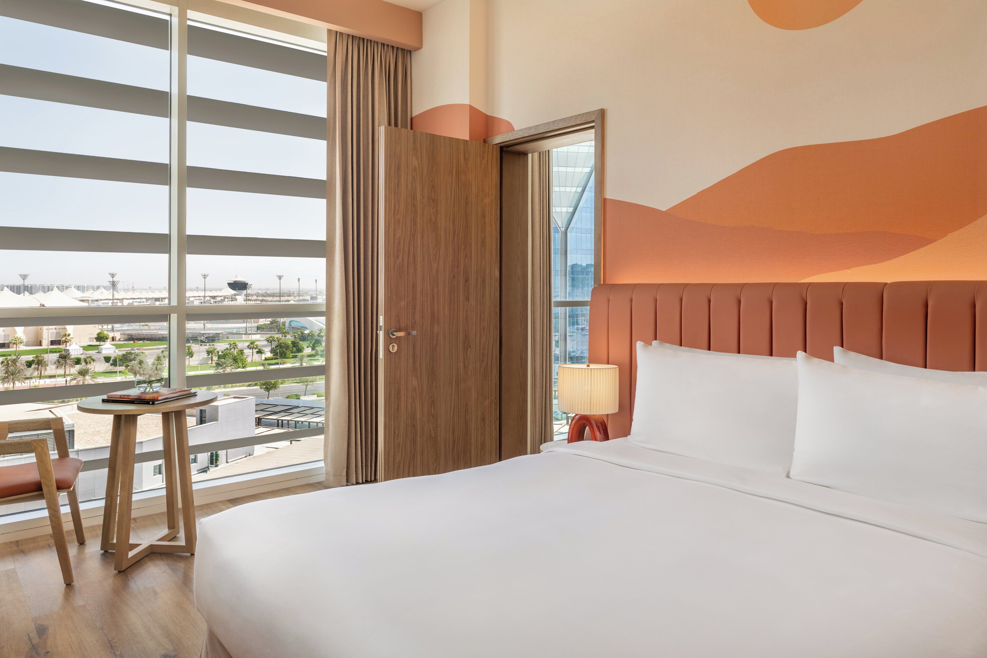 Une chambre d’hôtel avec un grand lit, une table de chevet en bois, une table avec une lampe et une vue sur la ville grâce à de grandes fenêtres du sol au plafond.