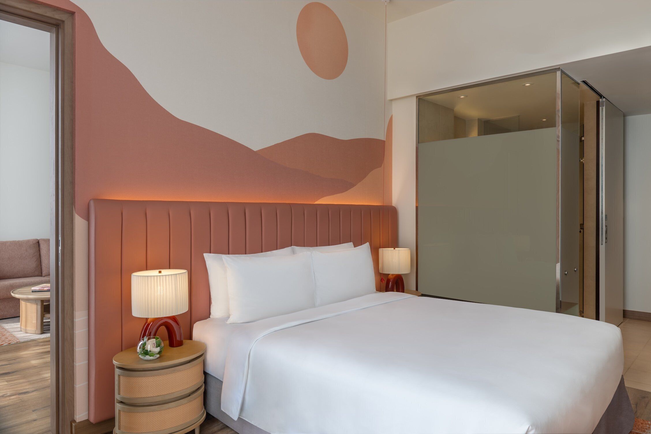 A hotel room with a large bed, a headboard with a unique design, two bedside lamps, a small table with a lamp, and a glass partition wall.