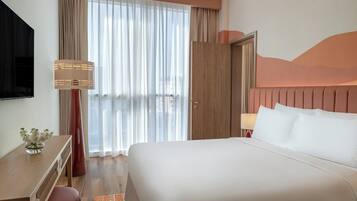 A hotel room with a large bed, a desk with a lamp, a TV, and a view of the city through sheer curtains.