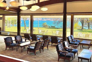 A spacious indoor area with large windows offering a view of a lake and trees. There are several wooden chairs and tables arranged for seating.