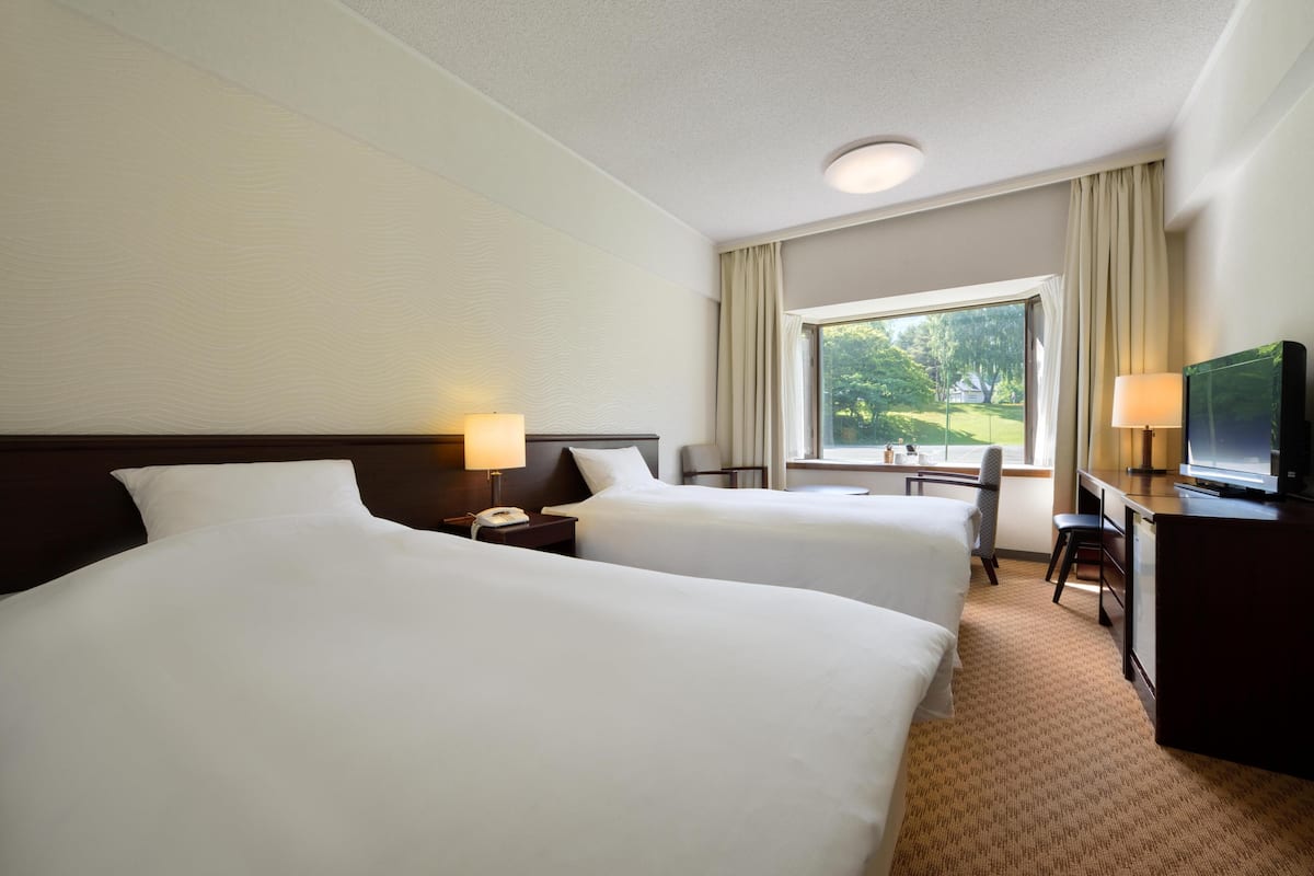 A hotel room with two beds, a desk, a television, and a window with a view of greenery.